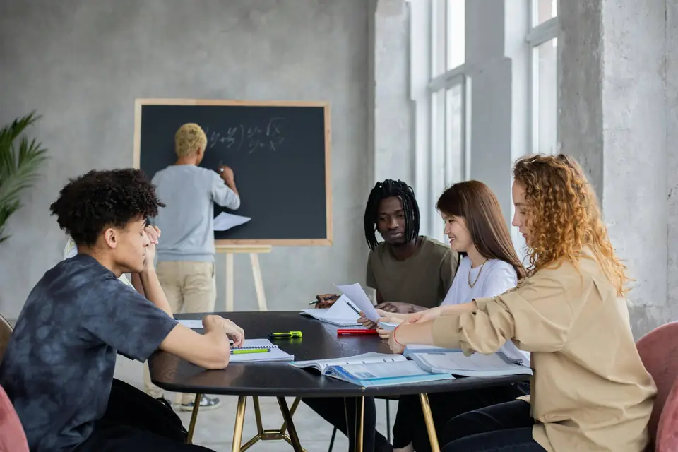 Alumnos trabajando alrededor de una mesa con apuntes y subrayadores mientras otro resuelve un ejercicio en la pizarra.